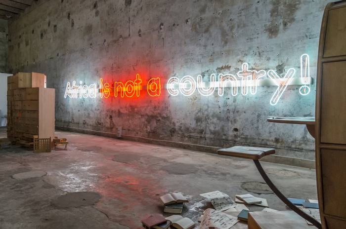 Exhibition room with a wooden archive structure, &laquo;Africa is not a country!&raquo; neon sign and books scattered on the floor