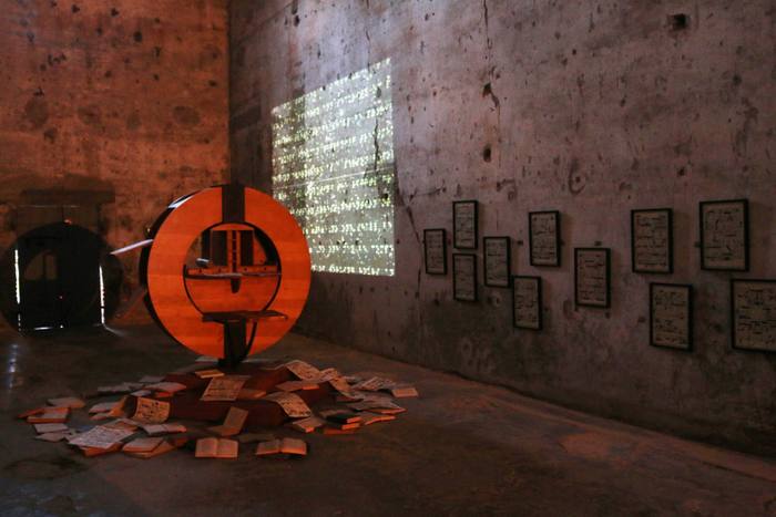 Orange circular wooden sculpture with books scattered on the floor and a projection of light dots on the wall