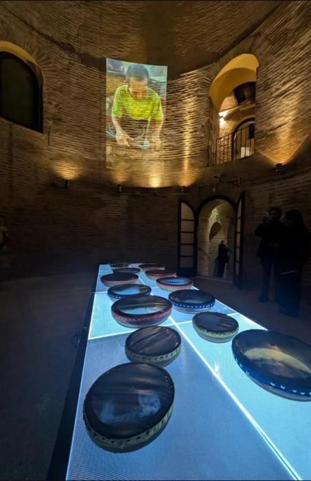 A long illuminated table with frame drums inside a brick tower, and a projected video on the wall