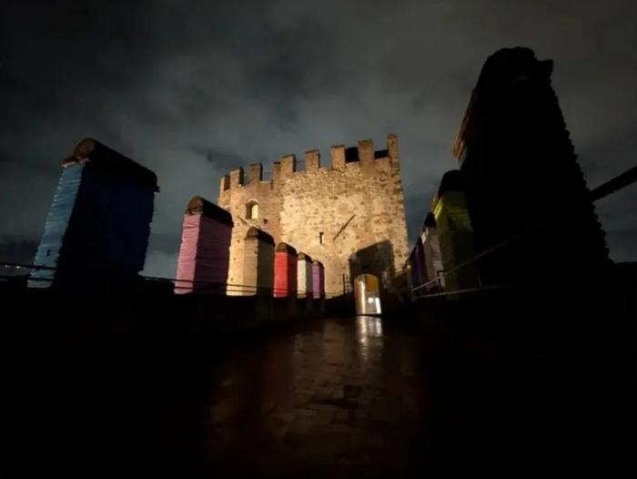 The covered walkway at night with stone columns wrapped in brightly coloured bands, leading towards a tower