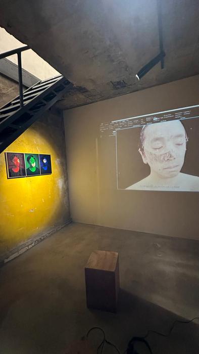 A video projection of a face beside a triptych of framed portraits on a yellow wall, under a staircase