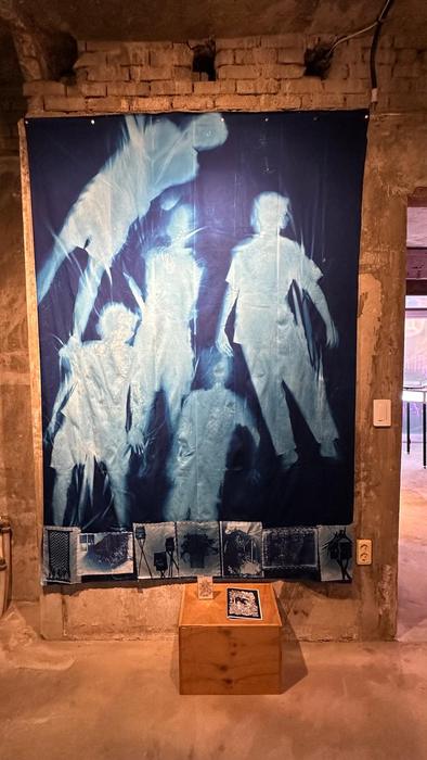 Large blue cyanotype textile showing silhouetted figures, with a wooden plinth below