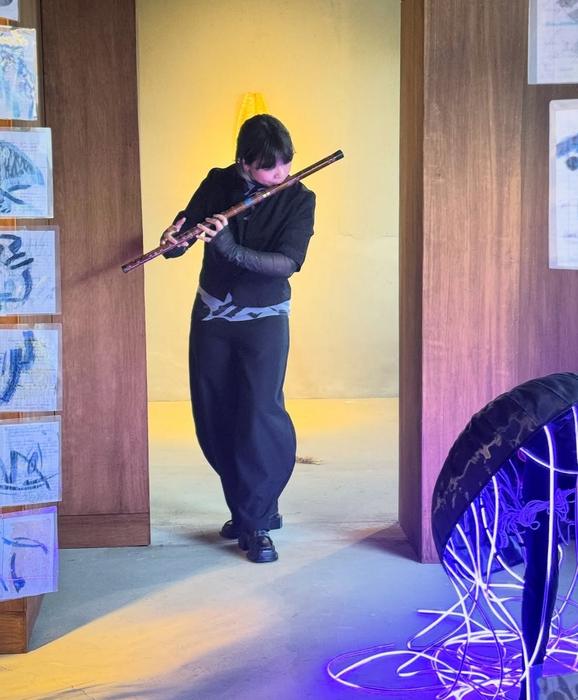 A performer playing a long flute in a doorway, among hanging drawings and a glowing installation