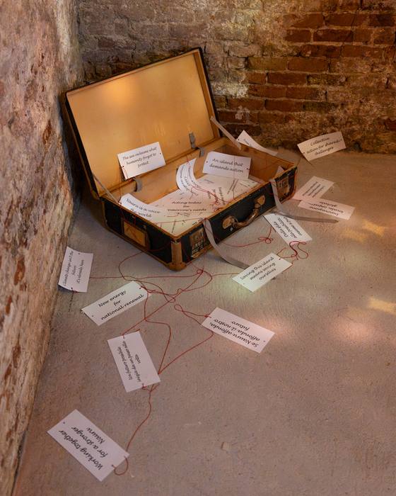 Open vintage suitcase installation with handwritten tags connected by red thread, on a brick floor
