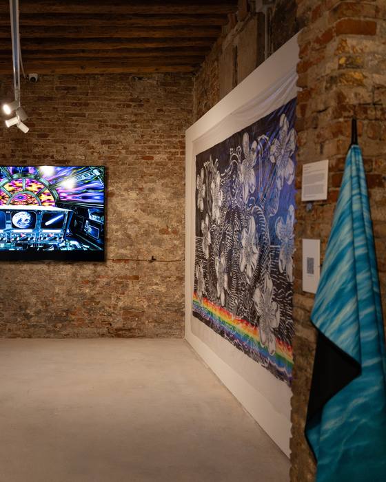 Video screen showing a colourful digital collage on a brick wall, next to a black-and-white floral textile and a turquoise fabric piece