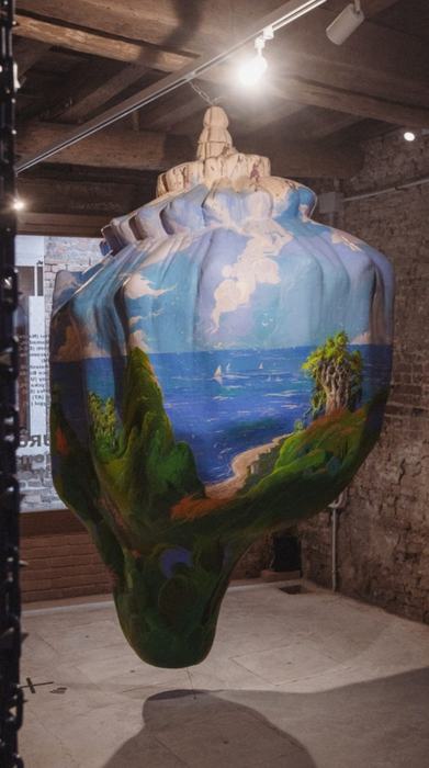 A large hanging sculpture shaped like a shell, painted with a sea and coastal landscape, in a brick room